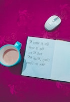 An open notebook with handwritten Hindi text is placed on a bright pink surface, surrounded by a white and silver wireless mouse and a teal cup containing a beverage. The background features a subtle floral pattern.