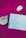 An open notebook with handwritten Hindi text is placed on a bright pink surface, surrounded by a white and silver wireless mouse and a teal cup containing a beverage. The background features a subtle floral pattern.