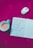 An open notebook with handwritten Hindi text is placed on a bright pink surface, surrounded by a white and silver wireless mouse and a teal cup containing a beverage. The background features a subtle floral pattern.
