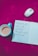 An open notebook with handwritten Hindi text is placed on a bright pink surface, surrounded by a white and silver wireless mouse and a teal cup containing a beverage. The background features a subtle floral pattern.