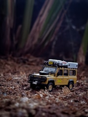 A detailed toy model of a classic off-road vehicle is placed on a natural, earthy surface resembling forest ground. The vehicle is equipped with roof racks and a spare tire on its hood, designed to resemble a rugged adventure-ready design.