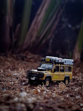 A detailed toy model of a classic off-road vehicle is placed on a natural, earthy surface resembling forest ground. The vehicle is equipped with roof racks and a spare tire on its hood, designed to resemble a rugged adventure-ready design.