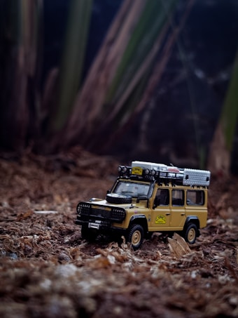 A detailed toy model of a classic off-road vehicle is placed on a natural, earthy surface resembling forest ground. The vehicle is equipped with roof racks and a spare tire on its hood, designed to resemble a rugged adventure-ready design.