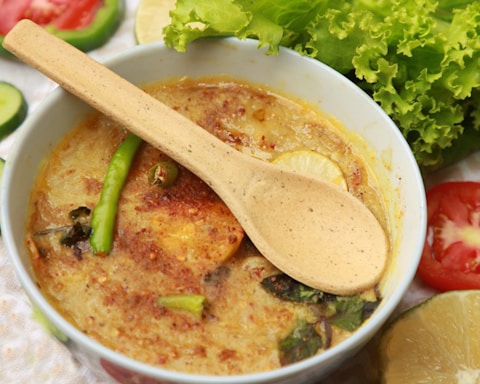 A bowl of spicy food featuring a creamy texture with various ingredients such as sliced green chilies, lemon slices, and herbs. A wooden spoon is placed across the bowl. Surrounding the bowl are fresh vegetables including lettuce, cucumber slices, tomato slices, and lemon wedges.