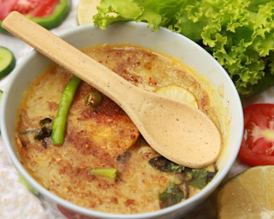 A bowl of spicy food featuring a creamy texture with various ingredients such as sliced green chilies, lemon slices, and herbs. A wooden spoon is placed across the bowl. Surrounding the bowl are fresh vegetables including lettuce, cucumber slices, tomato slices, and lemon wedges.
