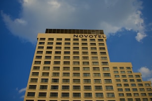 A tall, rectangular building with numerous evenly spaced windows stretches upward against a blue sky. The word 'NOVOTEL' is prominently displayed at the top of the hotel. Fluffy clouds are scattered across the sky.