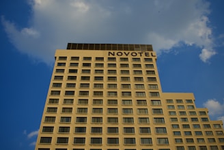 A tall, rectangular building with numerous evenly spaced windows stretches upward against a blue sky. The word 'NOVOTEL' is prominently displayed at the top of the hotel. Fluffy clouds are scattered across the sky.