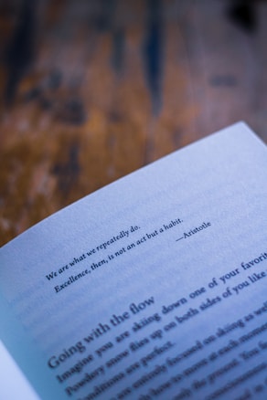 A close-up view of an open book displaying a quote by Aristotle, with blurred text visible at the bottom and a textured wooden surface in the background.