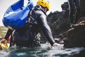 A person wearing a yellow helmet and a black wetsuit navigates through water while carrying a large blue bag. They are near rocky terrain, suggesting an outdoor adventure or coasteering activity.