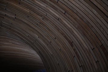 Curved wooden planks are arranged in a circular, tunnel-like structure. The planks feature a range of natural brown tones and are aligned in a seamless, repeating pattern.