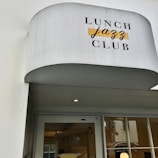 A white awning with the words 'Lunch Jazz Club' printed on it in contrasting fonts and colors. Below, there is a glass door with a reflection and a partial view of an interior space with yellow elements.