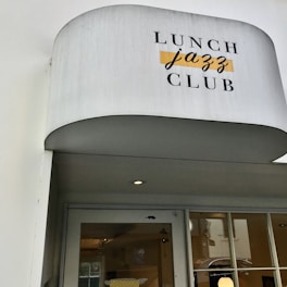 A white awning with the words 'Lunch Jazz Club' printed on it in contrasting fonts and colors. Below, there is a glass door with a reflection and a partial view of an interior space with yellow elements.