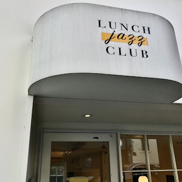 A white awning with the words 'Lunch Jazz Club' printed on it in contrasting fonts and colors. Below, there is a glass door with a reflection and a partial view of an interior space with yellow elements.