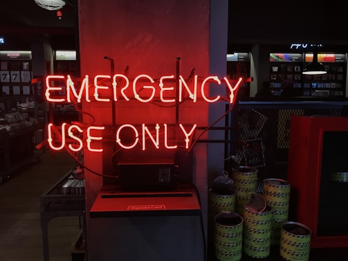 A neon red sign reading 'EMERGENCY USE ONLY' is prominently displayed on a wall. The surrounding environment appears to be dimly lit, with metal barrels labeled 'EMERGENCY' and some industrial-looking equipment in the background. Shelves and display counters are visible in a retail-like setting.