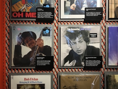 A collection of vinyl record covers displayed on a wall, featuring Bob Dylan albums. The covers include vibrant artwork and photographs of the musician. Each album cover has a small informational placard with details in Korean and English.