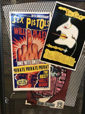 The image features three colorful concert posters attached to a metal mesh background. The top left poster is for the Sex Pistols' 30th anniversary, prominently displaying a hand with an extended middle finger. This poster has a blue, yellow, red, and white color scheme. The top right poster is for The Cure's European tour in 1979, showing an abstract drawing with a black background and yellow text. Below these, a third poster is partially visible with a vintage design, referencing The Fillmore venue, with a color scheme of pink and brown shades.