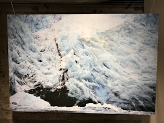 An image of a large, rugged glacier with distinct layers of ice and snow. The surface is uneven, with visible cracks and crevices. The glacier appears massive against a darker foreground, possibly a body of water.