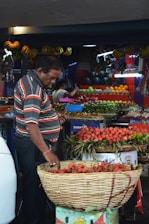 A man wearing a striped shirt is selecting lychees from a large basket at a fruit market stall. Various fruits such as bananas, oranges, and green apples are displayed on tables and hanging above. The space is filled with bright and colorful produce, creating a vibrant atmosphere.
