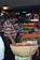A man wearing a striped shirt is selecting lychees from a large basket at a fruit market stall. Various fruits such as bananas, oranges, and green apples are displayed on tables and hanging above. The space is filled with bright and colorful produce, creating a vibrant atmosphere.