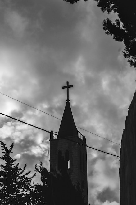 A tall stone bell tower with a cross on top is silhouetted against a cloudy sky. Telephone wires run across the image, and trees are visible at the bottom and along the edges.