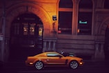 A vibrant orange sports car is parked in front of a historic stone building with ornate architectural details. The building has dark wooden doors and illuminated windows displaying signs for a financial and energy exchange group. There is a motorcycle with a fluorescent green seat cover parked nearby.