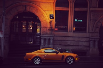 A vibrant orange sports car is parked in front of a historic stone building with ornate architectural details. The building has dark wooden doors and illuminated windows displaying signs for a financial and energy exchange group. There is a motorcycle with a fluorescent green seat cover parked nearby.
