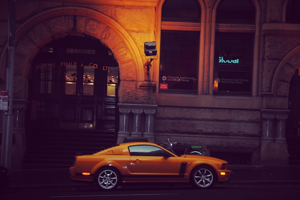 A vibrant orange sports car is parked in front of a historic stone building with ornate architectural details. The building has dark wooden doors and illuminated windows displaying signs for a financial and energy exchange group. There is a motorcycle with a fluorescent green seat cover parked nearby.