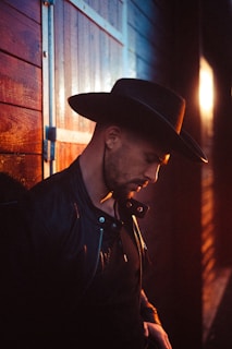 A person wearing a dark leather jacket and a wide-brimmed hat leans against a wooden wall. The lighting is warm, with a glow from the setting sun casting soft highlights and shadows.