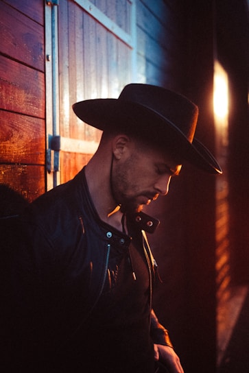 A person wearing a dark leather jacket and a wide-brimmed hat leans against a wooden wall. The lighting is warm, with a glow from the setting sun casting soft highlights and shadows.
