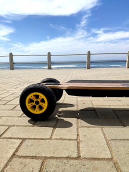 An electric skateboard with large yellow wheels is on a paved surface beside a seaside promenade. The ocean is visible in the background under a blue sky with some clouds.