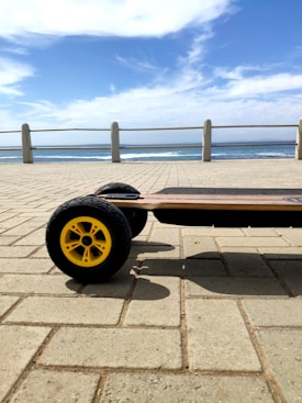 An electric skateboard with large yellow wheels is on a paved surface beside a seaside promenade. The ocean is visible in the background under a blue sky with some clouds.