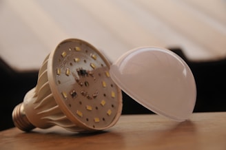 An LED light bulb is depicted with its cover removed, lying on a wooden surface. The internal components and LED chips are visible, showcasing its intricate design. The background is blurred, emphasizing the bulb as the primary subject.
