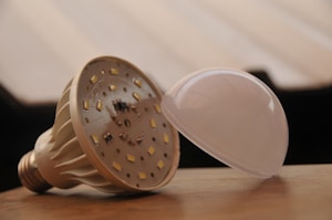 An LED light bulb is depicted with its cover removed, lying on a wooden surface. The internal components and LED chips are visible, showcasing its intricate design. The background is blurred, emphasizing the bulb as the primary subject.