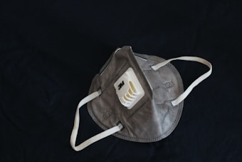 A grey protective face mask with white adjustable straps and a plastic valve on the front, placed on a black background. The mask displays the 3M logo and appears to be designed for respiratory protection.