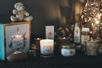 A cozy arrangement featuring a teddy bear, candles, and milk and honey-themed skincare products. The setting is accentuated with soft lighting from candles and a woven lamp, creating a warm, inviting atmosphere suitable for Christmas. Decorative elements like pine cones and rustic wooden candle holders enhance the festive theme.