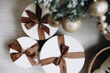 Three round gift boxes wrapped in white paper with brown ribbons are placed on a light wooden surface. Nearby, a decorated Christmas tree is visible with shimmering gold ornaments.
