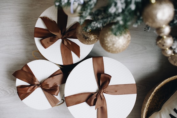 Three round gift boxes wrapped in white paper with brown ribbons are placed on a light wooden surface. Nearby, a decorated Christmas tree is visible with shimmering gold ornaments.