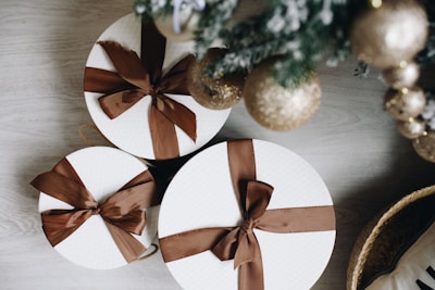 Three round gift boxes wrapped in white paper with brown ribbons are placed on a light wooden surface. Nearby, a decorated Christmas tree is visible with shimmering gold ornaments.