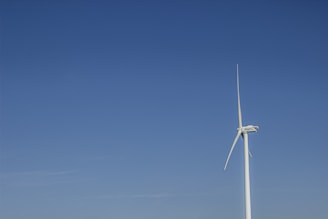 A large, white wind turbine stands against a clear blue sky, with no clouds visible. The turbine's blades are positioned to catch the wind, and its structure is sleek and modern.