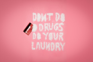 A pink background with a red credit card on the left. White powder spelling out 'DON'T DO DRUGS DO YOUR LAUNDRY' is arranged in the middle.