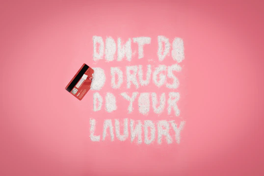 A pink background with a red credit card on the left. White powder spelling out 'DON'T DO DRUGS DO YOUR LAUNDRY' is arranged in the middle.