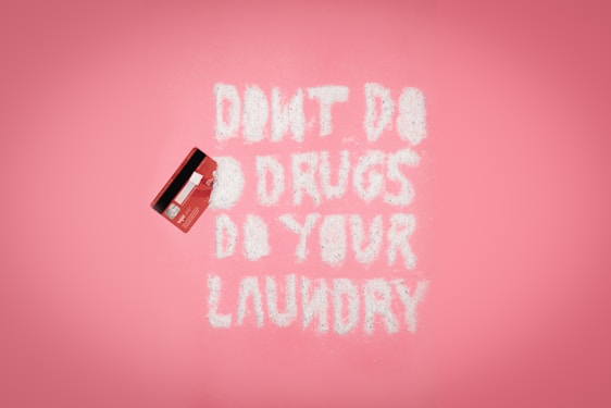 A pink background with a red credit card on the left. White powder spelling out 'DON'T DO DRUGS DO YOUR LAUNDRY' is arranged in the middle.