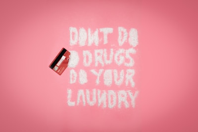 A pink background with a red credit card on the left. White powder spelling out 'DON'T DO DRUGS DO YOUR LAUNDRY' is arranged in the middle.