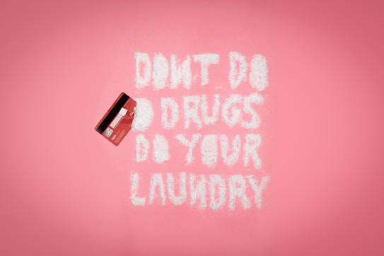A pink background with a red credit card on the left. White powder spelling out 'DON'T DO DRUGS DO YOUR LAUNDRY' is arranged in the middle.