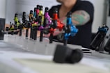 A series of colorful tattoo machines are displayed on a table, each mounted on a stand. The machines vary in color, including green, yellow, pink, red, and blue. In the background, the blurred image of a person with a tattooed arm is visible, suggesting a tattoo studio setting.