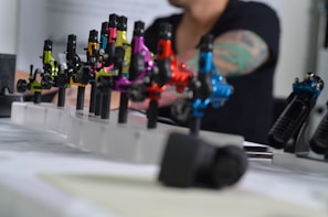 A series of colorful tattoo machines are displayed on a table, each mounted on a stand. The machines vary in color, including green, yellow, pink, red, and blue. In the background, the blurred image of a person with a tattooed arm is visible, suggesting a tattoo studio setting.