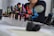 A series of colorful tattoo machines are displayed on a table, each mounted on a stand. The machines vary in color, including green, yellow, pink, red, and blue. In the background, the blurred image of a person with a tattooed arm is visible, suggesting a tattoo studio setting.