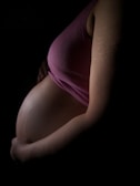 Side profile of a pregnant woman wearing a pink top, gently cradling her belly with one hand. The image is dimly lit, highlighting the curves and contours of the belly, with visible stretch marks.