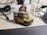 A detailed miniature model of an off-road vehicle, possibly a Land Rover, is placed on a desk. The model features roof racks with luggage and equipment, a spare tire on the hood, and various decals. Surrounding the vehicle are technological accessories such as cables and a microphone.