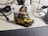 A detailed miniature model of an off-road vehicle, possibly a Land Rover, is placed on a desk. The model features roof racks with luggage and equipment, a spare tire on the hood, and various decals. Surrounding the vehicle are technological accessories such as cables and a microphone.